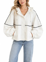 Woman wearing a cream oversized hooded jacket with black piping, front view with zipper closure and drawstring hood